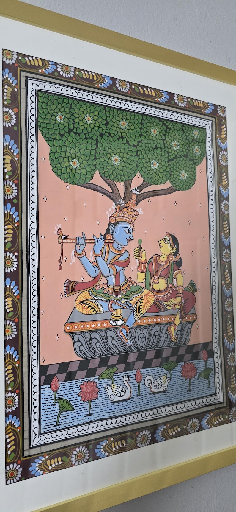 May include: A framed Indian painting depicting a couple under a tree. The figures are in traditional attire, with the male playing a flute. The artwork features vibrant colors, including blue, yellow, and red, and is set against a pink background.