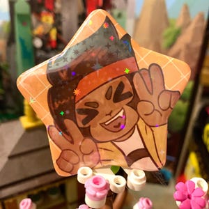 Könnte beinhalten: Ein holografischer sternförmiger Knopf mit einer Comicfigur mit brauner Haut, einem roten Stirnband und einer Friedensgeste. Die Figur lächelt und hat einen hellbraunen Hintergrund mit einem Gittermuster.