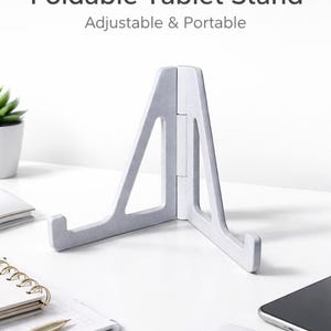 May include: A light gray foldable tablet stand. The stand is open and angled, with the text "Foldable Tablet Stand" and "Adjustable & Portable" above it. Other items on the white surface include a notebook, pen, and a smartphone.