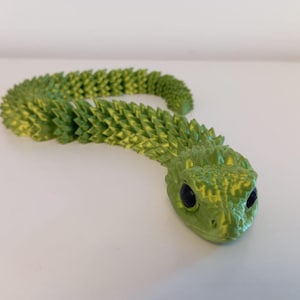 May include: A green and yellow articulated snake toy with black eyes. The snake is made of interlocking segments, allowing for flexible movement. The head is detailed with scales and the body has a textured pattern.