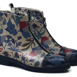 Könnte beinhalten: Knöchelstiefel mit floralem Muster. Die Stiefel haben einen beigefarbenen Hintergrund mit blauen, roten und grünen Blumenmustern. Sie haben eine schwarze Sohle, einen seitlichen Reißverschluss und eine Zuglasche hinten.