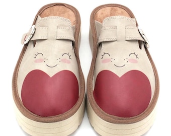 May include: Pair of beige clogs with a red heart design on the toe. The clogs have a smiling face design above the heart, a brown trim, and a buckle strap. The soles are a light beige color.
