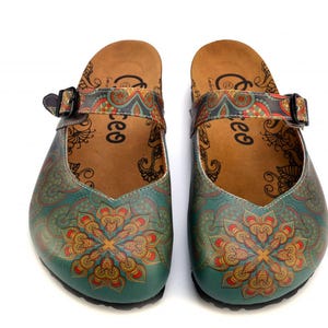 May include: A pair of teal and brown clogs with a floral design. The clogs have a strap with a buckle and a decorative pattern. The footbed is brown with a printed design and the word "ree" printed on the side.