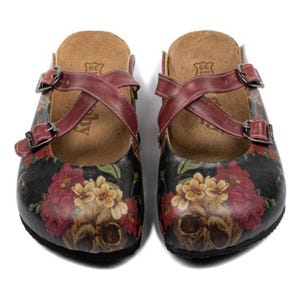 May include: Pair of black clogs featuring a floral and skull design. The clogs have burgundy leather straps and buckles. The footbed is light brown. The clogs are size 39.