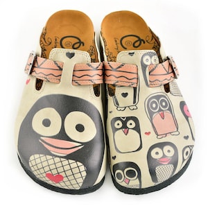May include: Cream-coloured clogs with penguin and owl designs. One clog features a large penguin face, while the other has multiple owl illustrations. Both have pink straps and buckles. The insoles are brown.