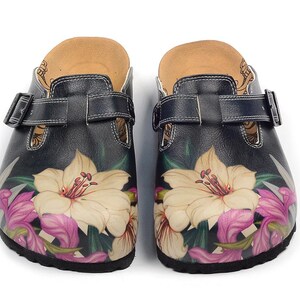 Könnte beinhalten: Ein Paar schwarze Clogs mit floralem Design. Die Clogs haben eine cremefarbene Blume mit rosa und grünen Akzenten. Die Clogs haben einen Schnallenriemen und ein Korkfußbett.