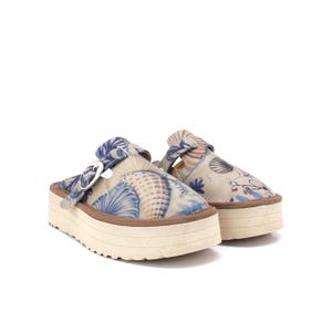 Könnte beinhalten: Paar Clogs mit Plateau und Muschelmuster in Blau und Beige. Die Clogs haben eine dicke, cremefarbene Sohle mit braunem Rand und einen Schnallenverschluss. Das Design hat ein nautisches Thema.