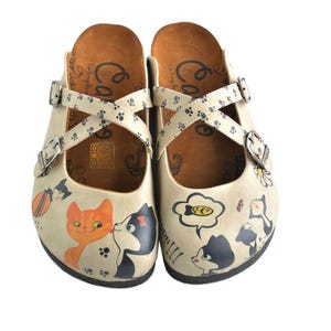 Könnte beinhalten: Cremefarbene Clogs mit Katzenillustrationen. Die Clogs haben gekreuzte Riemen mit Pfotenabdruck-Details und verstellbaren Schnallen. Die Katzendesigns zeigen orange, schwarze und weiße Katzen, Sprechblasen und florale Akzente.