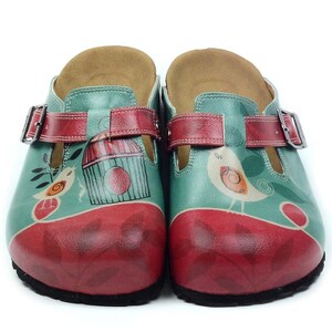 May include: A pair of teal and burgundy clogs with a bird and cage design. The clogs have a red leather strap and buckle closure. The toe area is burgundy with a leaf pattern. The clogs are a size 8.
