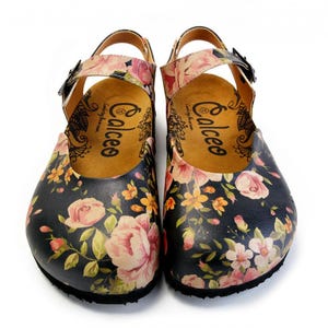 Pode incluir: Par de tamancos com padrão floral com base azul escuro, adornados com flores rosa e amarelas. Os tamancos têm uma tira e fecho de fivela. O interior é bege com a marca "Calceo".