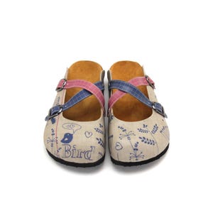 Könnte beinhalten: Paar beigefarbene Clogs mit Vogel- und Blumendesign. Die Clogs haben rosa und blaue gekreuzte Riemen mit silbernen Schnallen. Das Wort "Bird" ist auf der Seite aufgedruckt.