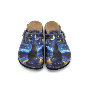 Puede incluir: Zuecos azules con un diseño de noche estrellada con una silueta de gato negro. Los zuecos tienen correas ajustables y un interior marrón. El diseño incluye una luna creciente y estrellas amarillas.