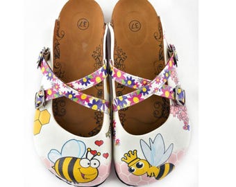 May include: Pair of white clogs with a bee theme. The clogs feature a cartoon bee design, floral straps, and a honeycomb pattern. The footbed is brown with a floral design, and the size is marked as 37.