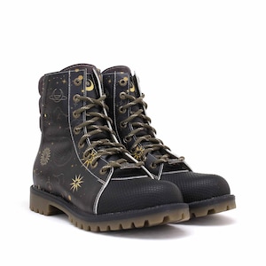 Op de afbeelding: Zwarte combat boots met een hemels ontwerp. De laarzen hebben gouden en witte accenten, waaronder sterren, manen en planeten. Ze hebben olijfgroene veters en een getextureerde neus. De zolen zijn lichtbruin.
