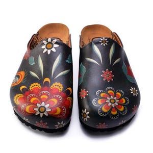 Peut inclure: Paire de sabots noirs ornés de motifs floraux colorés. Les sabots ont une semelle intérieure en liège et une semelle noire. Les motifs floraux comprennent des fleurs rouges, jaunes et blanches avec des feuilles vertes. Sabots à enfiler.