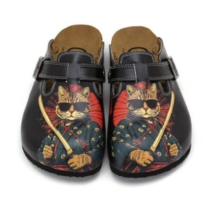 May include: Black clogs featuring a cat wearing sunglasses and a samurai outfit, holding a sword. The cat is set against a red and black background. The clogs have black straps and a brown footbed.