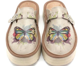 May include: A pair of beige platform clogs with a colorful butterfly design. The clogs have a brown trim, adjustable buckle straps, and a cream-colored sole. The butterfly is depicted with vibrant colors.
