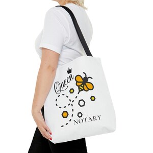 Puede incluir: Bolsa de tela blanca con asas negras y un diseño de abeja. La bolsa dice "Queen Notary" con una corona sobre la palabra "Queen".