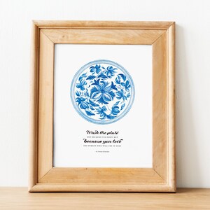 Op de afbeelding: Een ingelijst print met een blauw en wit bloemenontwerp op een bord. De tekst luidt "Was de plaat niet omdat hij vies is, maar omdat je van de persoon houdt die hem als volgende zal gebruiken."