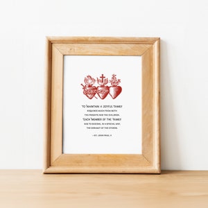 Op de afbeelding: Een ingelijste print met een rode illustratie van drie gestileerde harten met vlammen en een kruis. De tekst luidt: "Om een ​​vreugdevolle familie te behouden, is veel van zowel de ouders als de kinderen vereist. Elk lid van de familie moet op een speciale manier de dienaar van de anderen worden. -Johannes Paulus II"