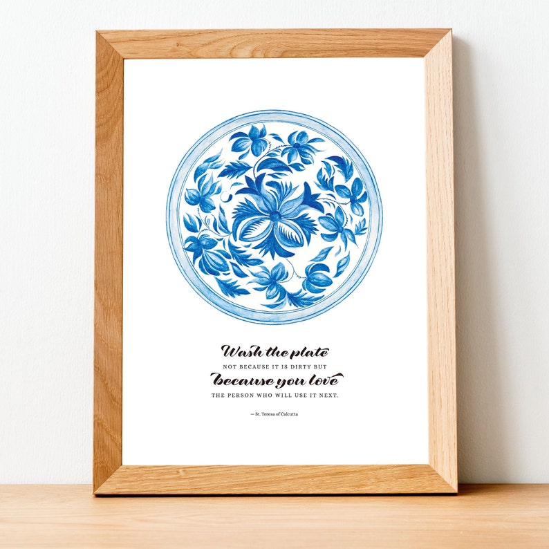 Op de afbeelding: Een ingelijst print met een blauw en wit bloemenmotief op een bord. De quote "Was de plaat niet omdat hij vies is, maar omdat je de persoon liefhebt die hem als volgende zal gebruiken." staat onder het bord gedrukt. De quote wordt toegeschreven aan St. Teresa van Calcutta.