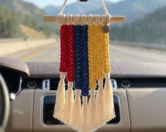 May include: A macrame car hanging ornament featuring vertical strands in red, navy blue, and yellow, suspended from a horizontal wooden dowel. The design includes a cream-colored cord for hanging and a series of cream-colored tassels at the bottom.