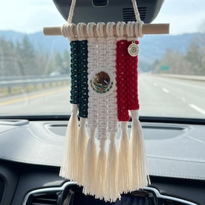 Könnte beinhalten: Ein Makramee-Autoanhänger in den Farben der mexikanischen Flagge aus dunkelgrüner, weißer und roter Baumwollkordel. Der mittlere weiße Bereich zeigt das mexikanische Wappen, und das Stück hängt an einem hölzernen Rundstab.