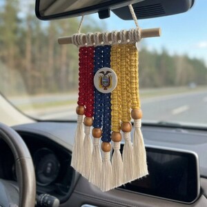 Könnte beinhalten: Ein Makramee-Autoanhänger mit vertikalen Kordeln in Rot, Blau und Gelb, den Farben der ecuadorianischen Flagge. Er enthält ein zentrales kreisförmiges Emblem des ecuadorianischen Wappens, Holzperlen und cremefarbene Fransenquasten als Abschluss.