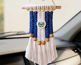 May include: A macrame car hanging ornament featuring alternating blue and white cotton cords suspended from a wooden dowel. The design includes a circular emblem of the El Salvador flag in the center and is accented with natural wooden beads.