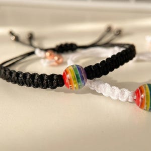 May include: Close-up of two friendship bracelets. One bracelet is black, the other is white. Each bracelet has a rainbow-colored glass bead in the center. The bracelets are on a white surface.