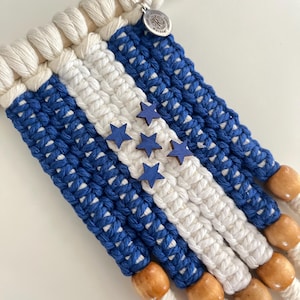 May include: A macrame wall hanging featuring a blue, white, and blue striped design with wooden star accents. The piece is crafted with white cotton rope and wooden beads, and is attached to a natural wooden dowel. A silver charm hangs from the dowel.