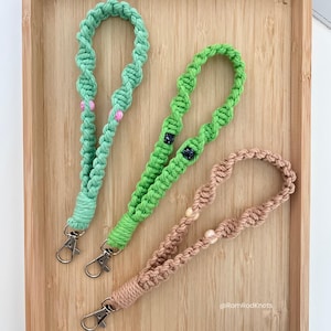 May include: Three macrame lanyards in shades of green and tan, each with a silver-colored clasp. The green lanyards have pink and black beads. The tan lanyard has light-colored beads. The lanyards are displayed on a wooden tray.