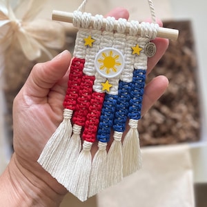 Könnte beinhalten: Ein kleiner Makramee-Wandbehang im Design der philippinischen Flagge aus cremefarbener, roter und blauer Baumwollschnur. Er enthält ein zentrales weißes kreisförmiges Emblem mit einer gelben Sonne, drei gelbe Holzsterne und einen Holzstab mit Aufhängeschnur.