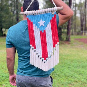 May include: A macrame wall hanging featuring the Puerto Rican flag design, crafted with light blue, white, and red cotton cord. The piece is suspended from a wooden dowel and finished with a series of white tassels along the bottom edge.