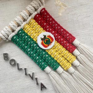 May include: A handmade macrame wall hanging featuring the colors of the Bolivian flag: red, yellow, and green. A white button with a colorful emblem is attached. The word "BOLIVIA" is spelled out in silver letters.