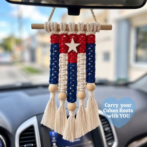 May include: A macrame car hanging ornament featuring the Cuban flag design in red, white, and blue cord. It includes a wooden dowel at the top, a central white star, natural wooden beads, and five tassels at the bottom. Text reads: Carry your Cuban Roots with YOU.