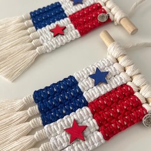 May include: Two macrame wall hangings with a flag design. Each features blue and red squares, white macrame, wooden dowels, and star accents. The design is reminiscent of a flag. The hangings are handmade and suitable for home decor.