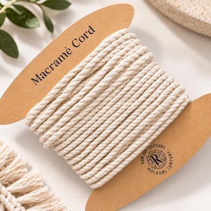 May include: A spool of off-white, twisted macramé cord wrapped around a brown cardboard holder. The holder features the text Macramé Cord at the top and a circular logo with the text RAM RBD CREATIONS MACRAMÉ COMPANY at the bottom right.