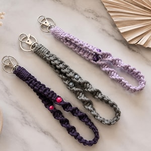 May include: Three macrame wristlet keychains featuring knotted cord in dark purple, gray, and light purple. Each keychain includes a silver-toned metal lobster clasp and two decorative beads integrated into the twisted cord design.