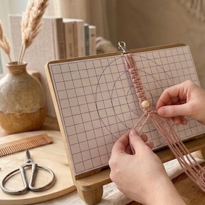 Puede incluir: Un tablero de macramé de madera con un patrón de cuadrícula y círculos concéntricos, utilizado para manualidades. Un proyecto de macramé rosa está en progreso, con una cuenta de madera y un cierre plateado. Tijeras y peine.