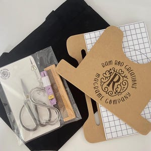 Puede incluir: Un kit de macramé con una bolsa de mano negra, tijeras, un peine y una cinta métrica. El kit incluye una plantilla de cartón con el texto "RAM ROD CREATIONS MACRAME COMPANY". Las tijeras están en un paquete de plástico transparente.