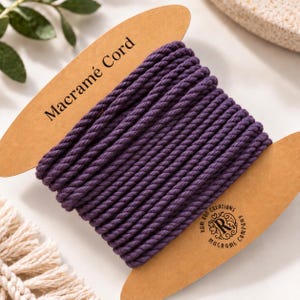 May include: A spool of deep purple twisted macramé cord wrapped around a tan cardboard holder. The holder features the text Macramé Cord at the top and a circular logo at the bottom right that reads Ram Rod Creations Macrame Company.