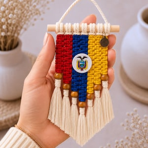 May include: A small macrame wall hanging featuring the yellow, blue, and red colors of the Ecuadorian flag. The design includes a central circular emblem of the Ecuadorian coat of arms, wooden beads, and off-white cotton tassels hanging from a wooden dowel.
