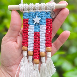 Könnte beinhalten: Ein kleiner Makramee-Wandbehang im Design der puerto-ricanischen Flagge aus roter, weißer und blauer Baumwollschnur. Er hängt an einem hellen Holzstab und ist mit einem silbernen Sternanhänger sowie Holzperlen am unteren Ende verziert.