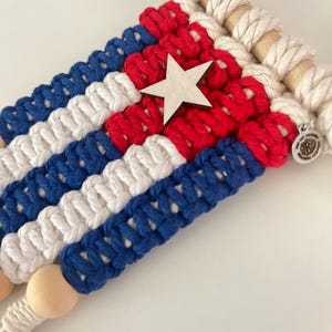 以下が含まれることがあります： 赤、白、青のデザインの、マクラメの壁掛け。中央に木の星があります。マクラメは木のダボに取り付けられており、下部に木製のビーズが付いています。小さな金属製のチャームが付いています。