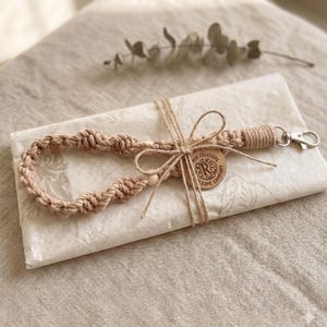 Könnte beinhalten: Ein handgefertigtes Makramee-Armband in hellem Braun, mit einem gewebten Design und einem silberfarbenen Verschluss. Das Armband ist mit Bindfaden und einem Holzanhänger mit der Aufschrift "ROD CREATIONS MACRAME COMPANY" gebunden.