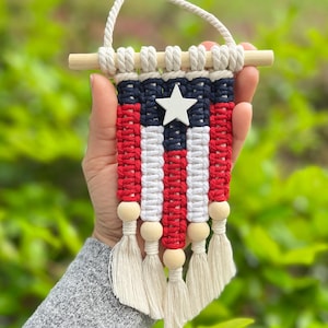 Könnte beinhalten: Ein kleiner Makramee-Wandbehang mit einem Muster aus roter, weißer und marineblauer Kordel. Das Design umfasst einen zentralen weißen Holzstern, einen horizontalen Holzstab an der Oberseite mit einer Schlaufe sowie fünf Quasten mit Holzperlen.