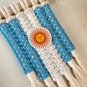 Peut inclure: Décoration murale en macramé représentant le drapeau argentin. Elle présente des rayures bleu clair et blanches, avec un emblème central du soleil. Le macramé est fixé à une cheville en bois.