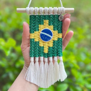Könnte beinhalten: Ein Makramee-Wandbehang mit grünem und gelbem Garn in geometrischem Muster, aufgehängt an einem hellen Holzstab mit weißem Seil. Ein kreisförmiges blaues Emblem mit weißen Sternen und dem Text "ORDEM E PROGRESSO" ist in der Mitte platziert.