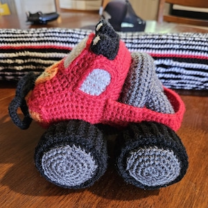 Puede incluir: Un camión de juguete de crochet rojo con ruedas negras y neumáticos de crochet grises. El camión tiene un corazón blanco de crochet en la parte delantera.